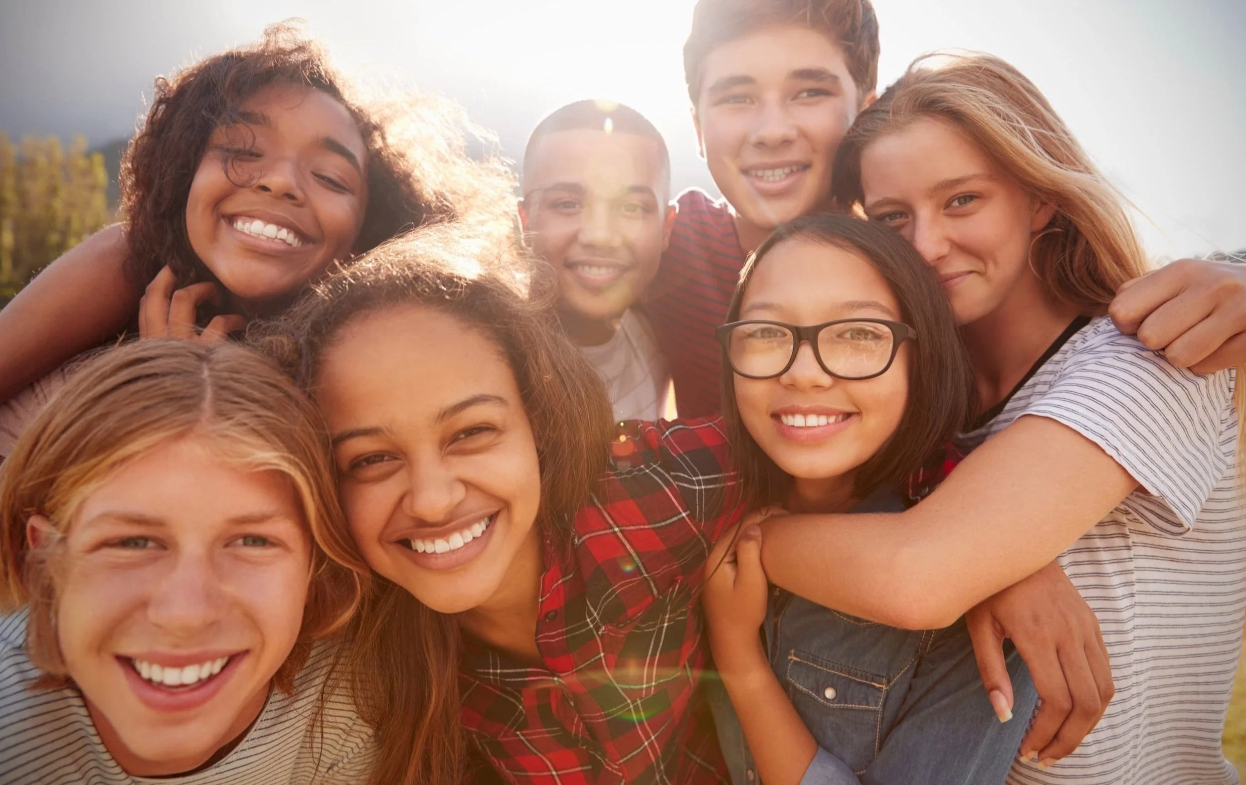 A group of seven smiling teenagers outdoors with their arms around each other in the sunlight